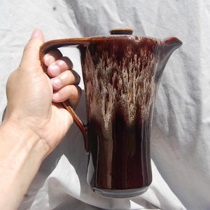 May include: A tall, brown ceramic coffee pot with a lid and a large handle. The pot has a glossy finish with a white and brown pattern. The coffee pot is held by a hand.