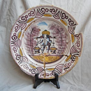 May include: An antique ceramic plate with a hand-painted scene of a person carrying baskets, set against trees and a blue sky. The plate has a decorative border with purple, yellow, and white patterns. The plate is cracked and displayed on a black stand.