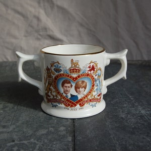 Puede incluir: Taza de cerámica blanca con dos asas, con un diseño conmemorativo de la boda del príncipe Carlos y Lady Diana Spencer. El diseño incluye sus retratos, escudos de armas y el texto "To Commemorate Their Marriage 29th July 1981."
