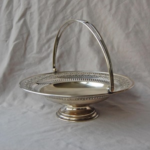 May include: Silver-plated serving basket with a handle. The basket has a round, shallow bowl with a decorative pierced border. The handle arches over the bowl and is attached on either side. The basket sits on a pedestal base.