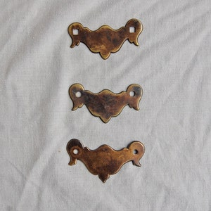 Può includere: Tre tiretti per cassetti in ottone antico con disegni decorativi. Ogni tiretto ha una forma rettangolare con la parte superiore e inferiore arrotondata. I tiretti sono in uno stato usurato e invecchiato, il che conferisce loro un aspetto vintage.