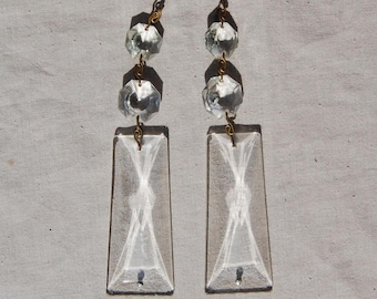 Puede incluir: Dos prismas de araña de cristal transparente con conectores de color latón. Cada prisma tiene forma rectangular con un diseño decorativo en el centro y tres cuentas de cristal facetadas encima.
