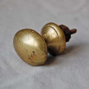 May include: Two antique brass door knobs with an oval shape. The knobs have a warm, golden hue and show signs of age and wear. The hardware is likely for a cabinet or door.