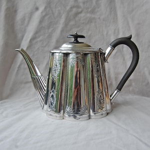 May include: A silver teapot with a black handle and lid. The teapot features a fluted body with etched decorative patterns. The spout is long and slender, and the handle curves elegantly. The lid has a small, round black knob.