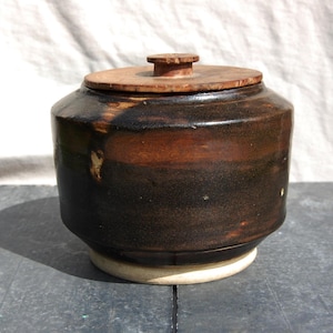 May include: A brown ceramic jar with a wooden lid. The jar has a dark brown glaze with a light brown stripe around the middle. The lid is made of wood and has a knob on top.