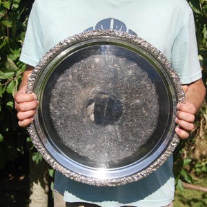 Puede incluir: Una bandeja redonda grande de plata con un diseño ornamentado y en relieve. La bandeja tiene un borde elevado y una superficie lisa y reflectante.