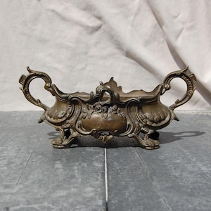 Könnte beinhalten: Ein längliches Gefäß aus bronzefarbenem Metall mit zwei verzierten, geschwungenen Griffen an den Seiten. Der Mittelteil zeigt detaillierte Reliefs mit Blumenmotiven und Schnörkeln und wird von vier kleinen, dekorativen Füßen an der Basis getragen.