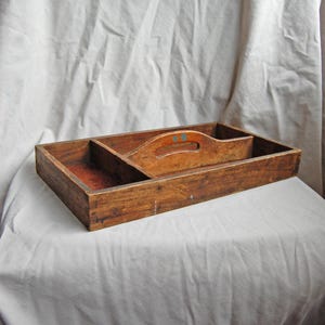 Op de afbeelding: Een vintage houten gereedschapsbak met een handvat en meerdere compartimenten. De bak is gemaakt van verouderd hout met zichtbare nerf en een natuurlijke afwerking. Het handvat heeft blauwe en groene accenten.