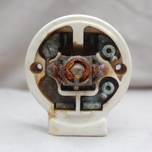 Può includere: Primo piano di un componente elettrico vintage bianco sporco. L'interno rivela componenti metallici con segni di ruggine e usura. Una vite centrale e l'hardware circostante sono visibili all'interno dell'alloggiamento circolare. Il componente ha una base rettangolare.