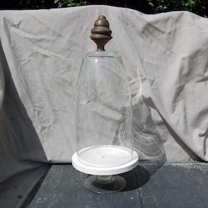 May include: A clear glass cloche with a rounded top and a metallic, acorn-shaped finial. The cloche rests on a circular white base, which is supported by a clear glass pedestal stand, creating a tall, vertical display vessel for objects.
