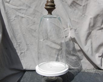 May include: A clear glass cloche with a rounded top and a metallic, acorn-shaped finial. The cloche rests on a circular white base, which is supported by a clear glass pedestal stand, creating a tall, vertical display vessel for objects.