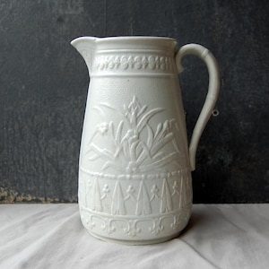 Op de afbeelding: Witte keramische kan met een handvat en tuit. De kan heeft reliëf bloemen- en geometrische patronen. De kan is een klassiek ontwerp, geschikt voor het serveren van water of andere dranken. De kan is waarschijnlijk vintage of antiek.