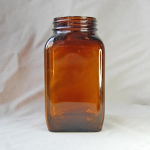 Puede incluir: Un tarro de vidrio ámbar vintage con un cuerpo cuadrado y un cuello roscado. El tarro tiene un color marrón cálido y rico y probablemente se utiliza para el almacenamiento. El vidrio es ligeramente reflectante y el tarro parece estar en buenas condiciones.