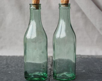 May include: Two green glass bottles with cork stoppers. The bottles have a classic shape with a narrow neck and a wider base. The corks are light brown. The bottles are displayed on a dark grey surface.