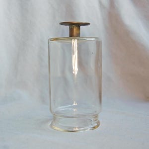 Peut inclure: Une bouteille en verre transparent avec un corps cylindrique et un bouchon de couleur laiton. La bouteille a une base légèrement évasée et un design simple. Le bouchon est en forme de disque plat. La bouteille est vide et semble vintage.
