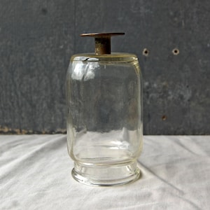 May include: A clear glass jar with a cylindrical body and a flared base. It has a small, round, bronze-coloured lid and handle. The jar is empty and sits on a white surface against a grey background.