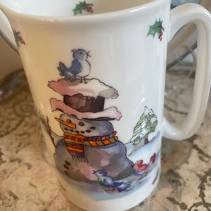 May include: White ceramic jug with a winter scene featuring a snowman with a colourful scarf, a carrot nose, and a top hat. Bluebirds and holly sprigs decorate the jug. The handle is square.