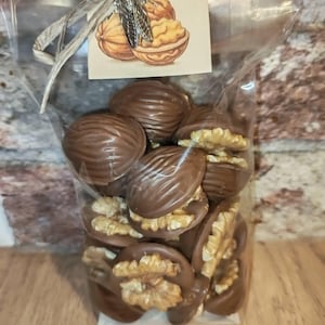 Peut inclure: Un sachet en plastique transparent rempli de moitiés de noix enrobées de chocolat. Le sachet est noué avec une ficelle grise et une étiquette carrée illustrée de noix, avec une breloque en métal argenté en forme de cigale attachée.