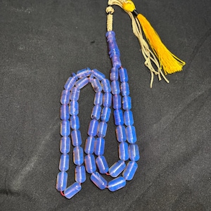 Puede incluir: Un collar de cuentas de oración azul con una borla amarilla. Las cuentas son de un azul translúcido con forma cilíndrica. La borla es amarilla y negra, unida a un cordón beige. El collar se muestra sobre una superficie negra.