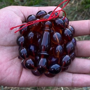 Puede incluir: Un juego de cuentas de oración de color ámbar oscuro, sostenido en una mano. Las cuentas son redondas y lisas, con una cuenta central alargada. Una cuerda roja está unida en la parte superior. El fondo es verde borroso.