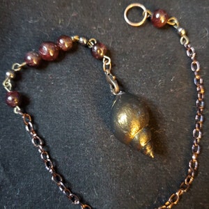 May include: A gold-colored shell pendant hangs from a chain with dark red beads. The chain is made of copper and silver.