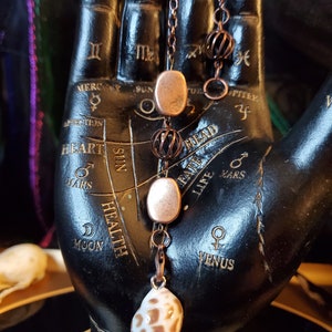 May include: A copper chain necklace with three copper beads and a seashell pendant. The necklace is displayed on a black hand with a palm reading chart.