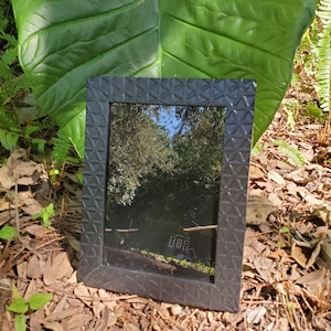 May include: A black picture frame with a geometric pattern on the border. The frame is reflecting a view of trees and a building in the background.
