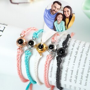 May include: Five colourful braided bracelets with eye-shaped charms. The charms are silver, gold, and black. The bracelets are on a white surface with a book in the background. The book has the word "BAKE" on the cover.