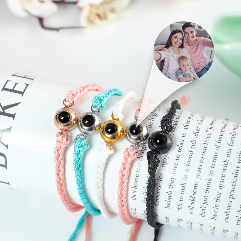 Puede incluir: Cinco pulseras trenzadas de colores vivos con un peque&ntilde;o dije redondo en el centro de cada una. Los dijes son plateados, dorados y negros. Las pulseras est&aacute;n sobre una superficie blanca con texto en el fondo.
