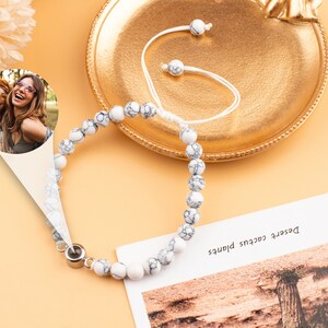 Puede incluir: Pulsera de cuentas de m&aacute;rmol blanco con un dije de plata. La pulsera est&aacute; sobre una superficie naranja claro con una foto de dos mujeres en el fondo.