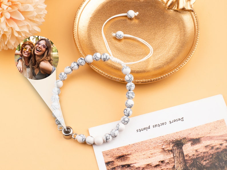 Puede incluir: Pulsera de cuentas de m&aacute;rmol blanco con un dije de plata. La pulsera est&aacute; sobre una superficie naranja claro con una foto de dos mujeres en el fondo.