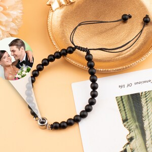 Puede incluir: Un brazalete de cuentas negras con un peque&ntilde;o dije de plata. El dije tiene una foto de una pareja en un entorno de boda. El brazalete est&aacute; sobre una superficie marr&oacute;n claro.