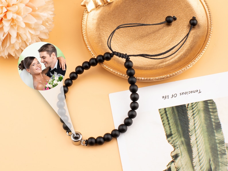 Puede incluir: Un brazalete de cuentas negras con un peque&ntilde;o dije de plata. El dije tiene una foto de una pareja en un entorno de boda. El brazalete est&aacute; sobre una superficie marr&oacute;n claro.