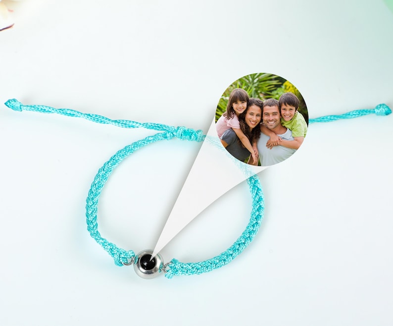Puede incluir: Un brazalete trenzado turquesa con una cuenta de plata que contiene una foto negra. La foto muestra a una familia de cuatro personas sonriendo en un parque.