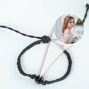 Puede incluir: Un brazalete trenzado negro con una cuenta negra. El brazalete est&aacute; atado con un nudo y tiene una peque&ntilde;a foto de una mujer con un vestido blanco.
