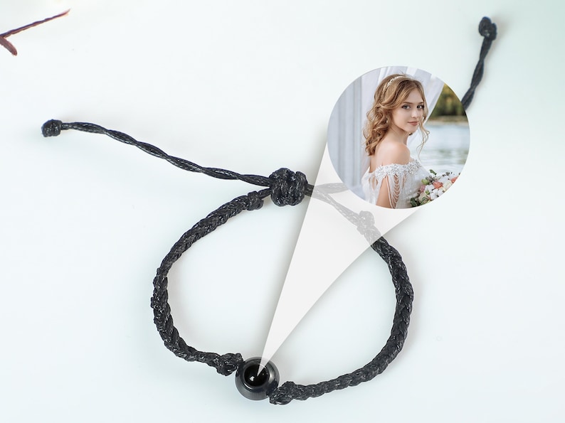 Puede incluir: Un brazalete trenzado negro con una cuenta negra. El brazalete est&aacute; atado con un nudo y tiene una peque&ntilde;a foto de una mujer con un vestido blanco.