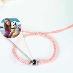 Puede incluir: Un brazalete trenzado rosa con un dije redondo de plata que presenta una foto de una mujer y su perro. La foto muestra a una mujer con cabello largo y casta&ntilde;o que lleva una camisa azul y blanca, sosteniendo un perro blanco con manchas marrones.