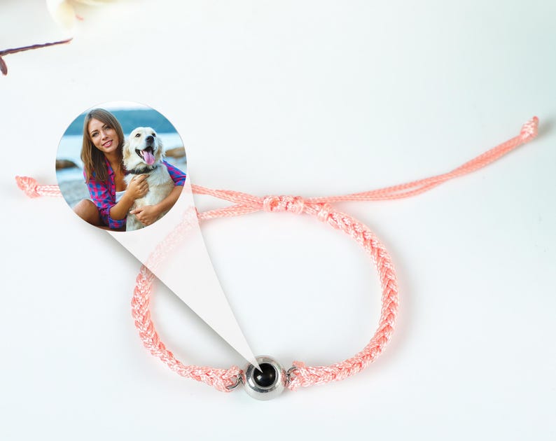Puede incluir: Un brazalete trenzado rosa con un dije redondo de plata que presenta una foto de una mujer y su perro. La foto muestra a una mujer con cabello largo y casta&ntilde;o que lleva una camisa azul y blanca, sosteniendo un perro blanco con manchas marrones.