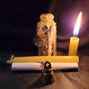 May include: A small glass bottle filled with herbs, crystals, and a pentagram charm. The bottle is sealed with wax and has a string tied around it. A white candle and a brass bell are also in the image.