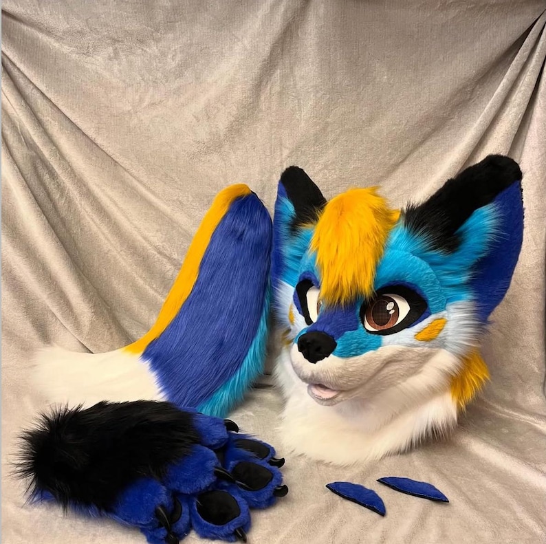 May include: A blue and yellow furry animal head with black ears and a white muzzle. The head is part of a full-body costume, which also includes a blue and yellow tail, blue and black paws, and two small blue ear pieces.