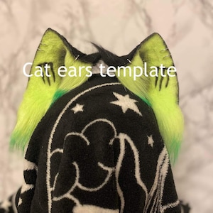 Op de afbeelding: Een paar groene kattenoren met zwarte stiksels en een witte basis. De oren zijn vastgemaakt aan een zwarte stof met een wit sterrenpatroon. De tekst "Cat ears template" is in wit op de stof geschreven.