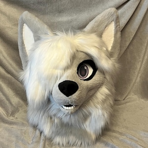 May include: A gray and white wolf fursuit head with a white streak of fur on the top of the head. The fursuit has a black nose and pink eyes.