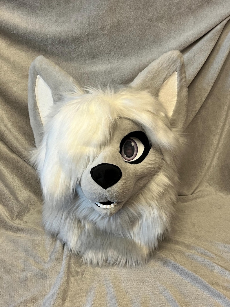 May include: A gray and white wolf fursuit head with a white streak of fur on the top of the head. The fursuit has a black nose and pink eyes.