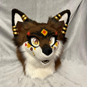 May include: A brown and white furry animal head with gold accents. The head has a black nose and yellow eyes. The fur is soft and fluffy. The head is a costume piece.