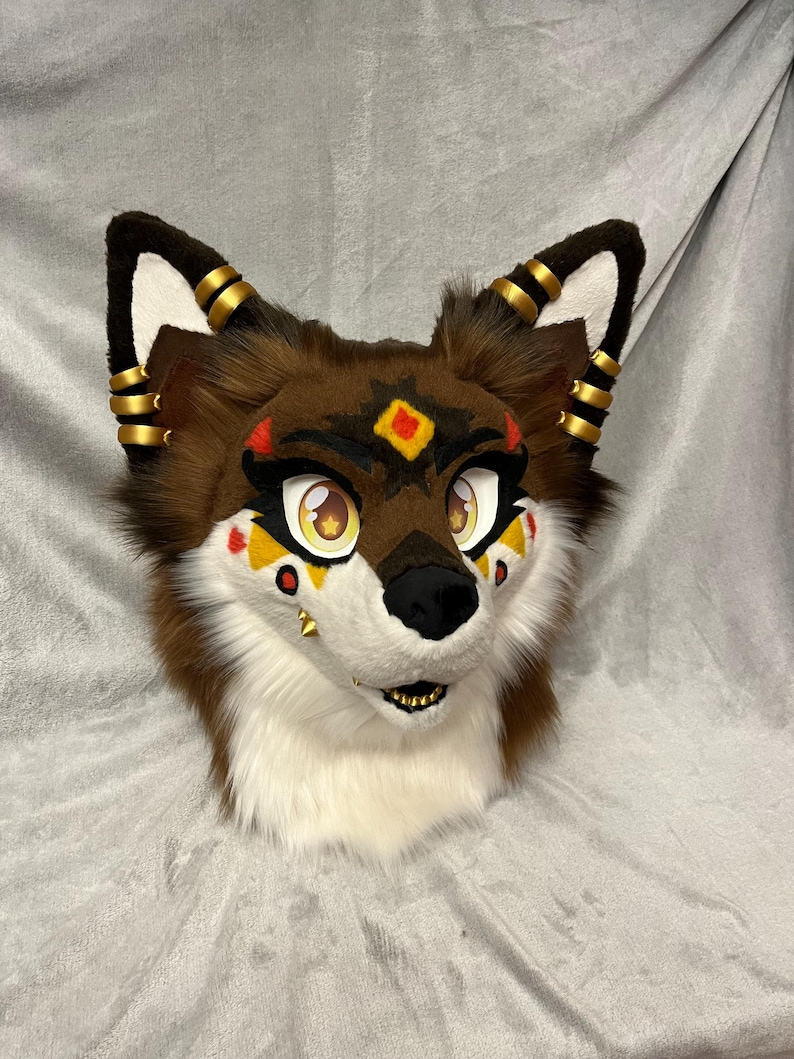 May include: A brown and white furry animal head with gold accents. The head has a black nose and yellow eyes. The fur is soft and fluffy. The head is a costume piece.