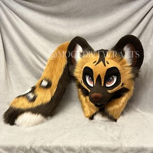 May include: A handmade African wild dog fursuit head and tail set. The head features a tan and brown color scheme with black accents around the eyes and ears. The tail is long and fluffy, with a similar color pattern.
