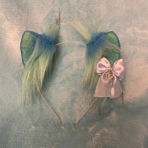 May include: A headband with two fluffy green and blue cat ears and a lavender bow with a gold heart charm.