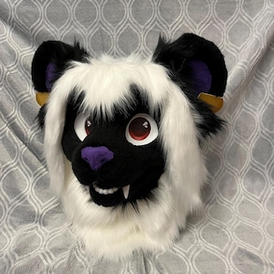 May include: A white and black furry animal head with red eyes and purple nose. The head has long white fur and black ears with purple accents. The head is on a gray patterned background.