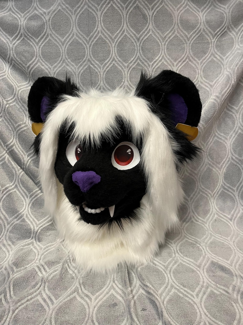 May include: A white and black furry animal head with red eyes and purple nose. The head has long white fur and black ears with purple accents. The head is on a gray patterned background.