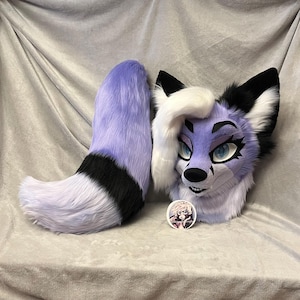 May include: A lavender and white furry fox head and tail set. The fox head has blue eyes, black accents, and white hair. The tail is lavender with a black stripe and white tip. A small round pin with a graphic is next to the head.